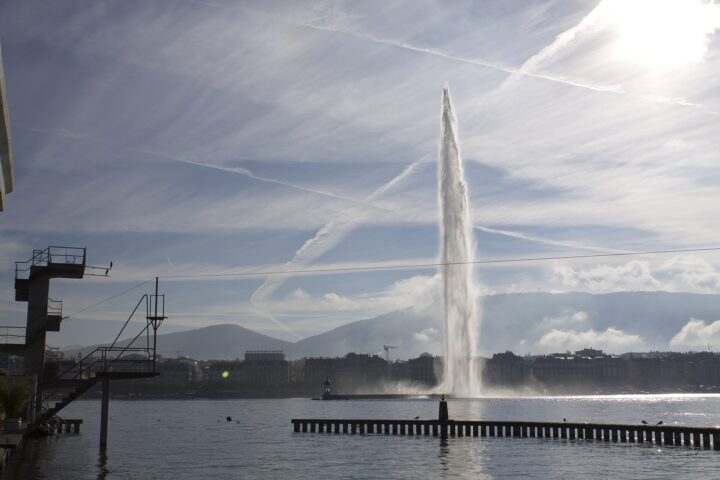 The Jet d'Eau rising against a streaked blue sky across Geneva's harbour, seen from the Bains des Pâquis with part of the diving tower on the left