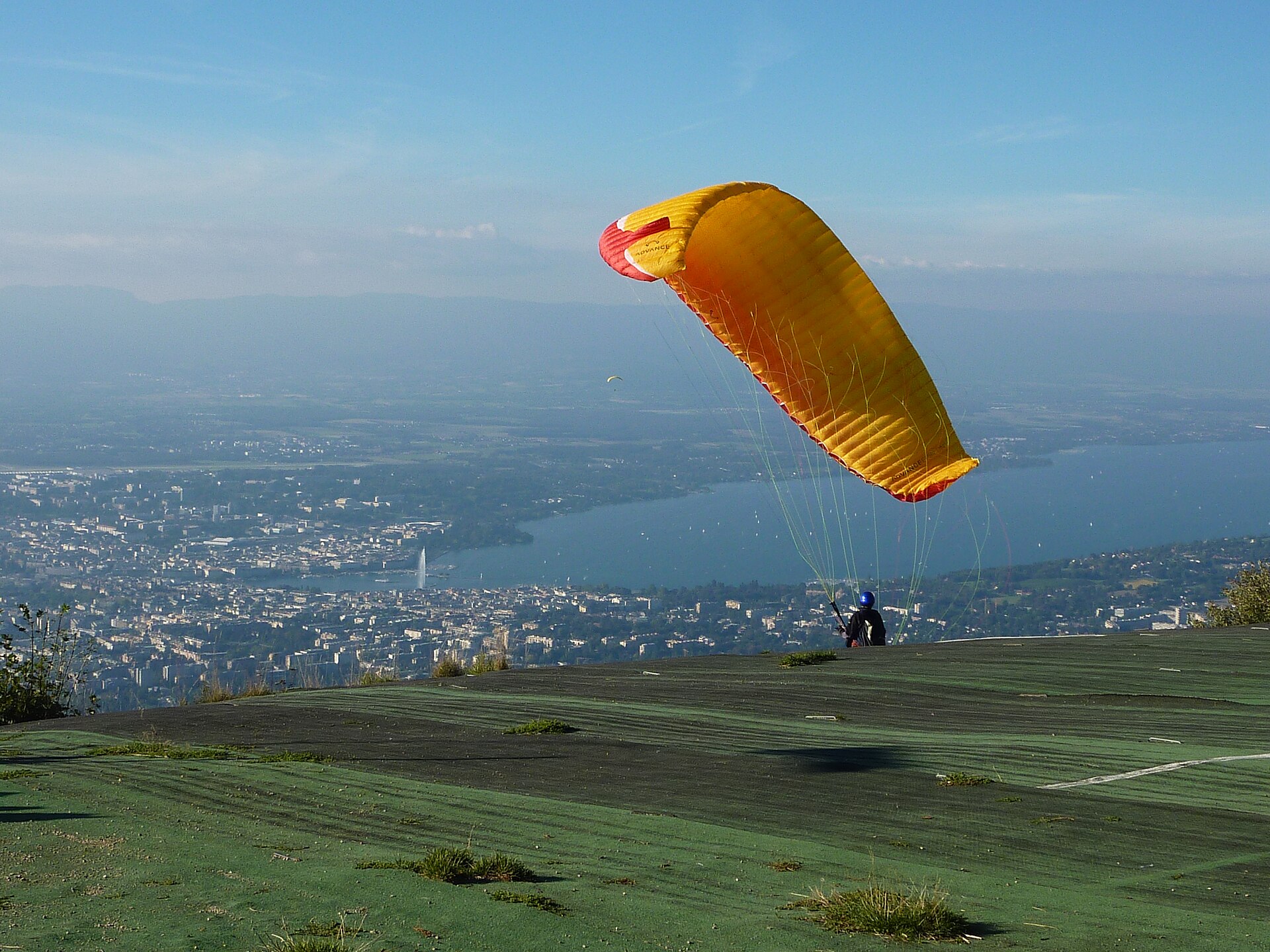 A paraglider with a yellow wing taking off from the launch area on the Salève, with Geneva and the lake below in the haze