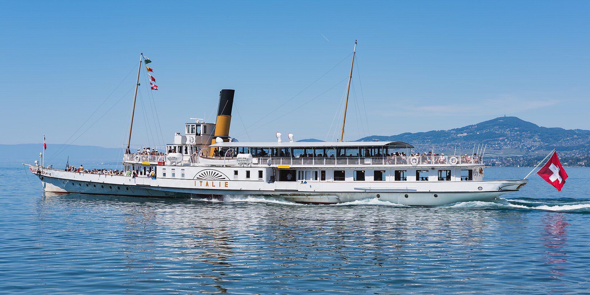 The white Belle Époque paddle steamer Italie with a yellow funnel sailing on Lake Geneva under a blue sky, a Swiss flag at the stern