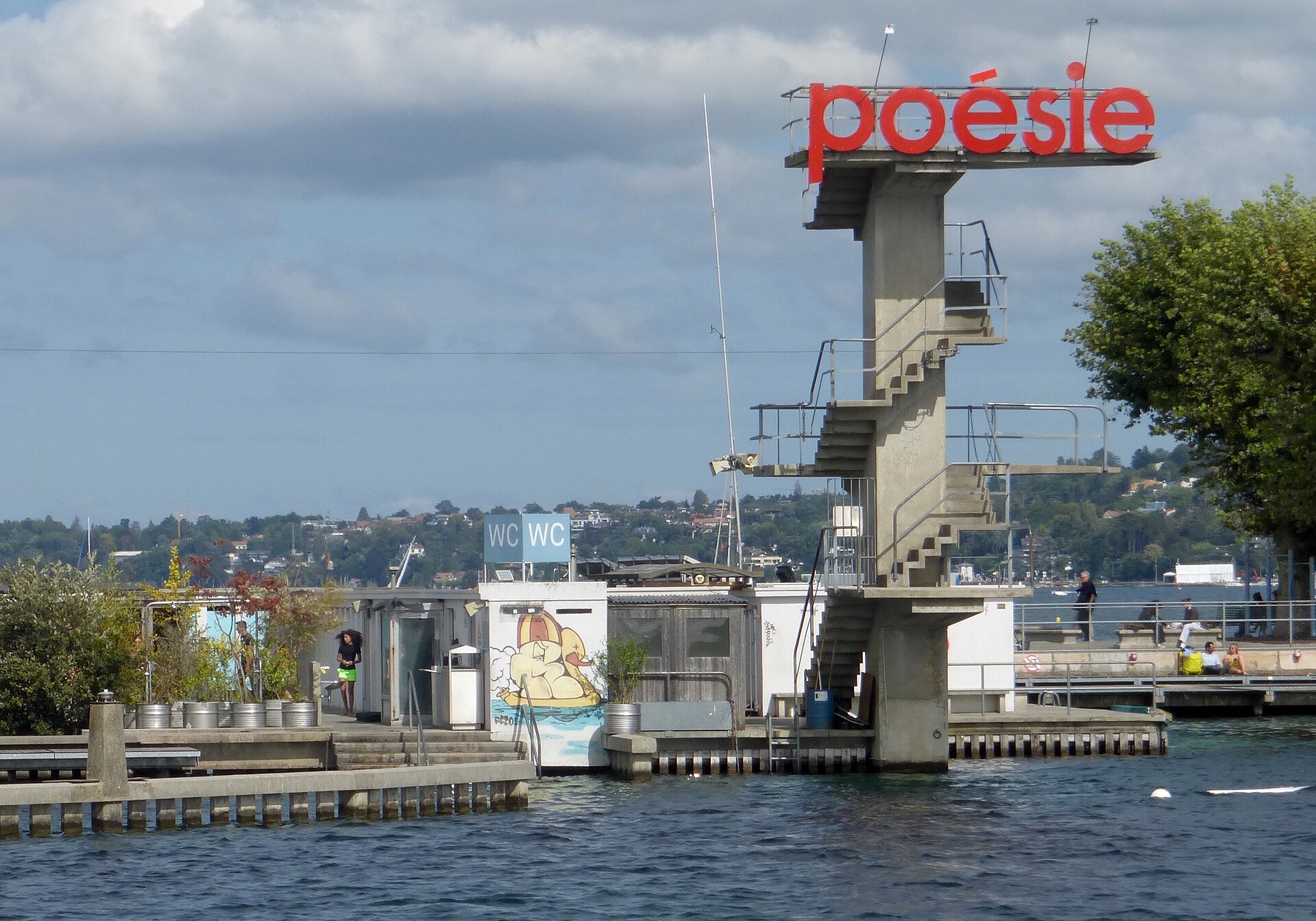 The concrete diving tower at the Bains des Pâquis in Geneva, with red lettering on top and the lake and the city's shore behind