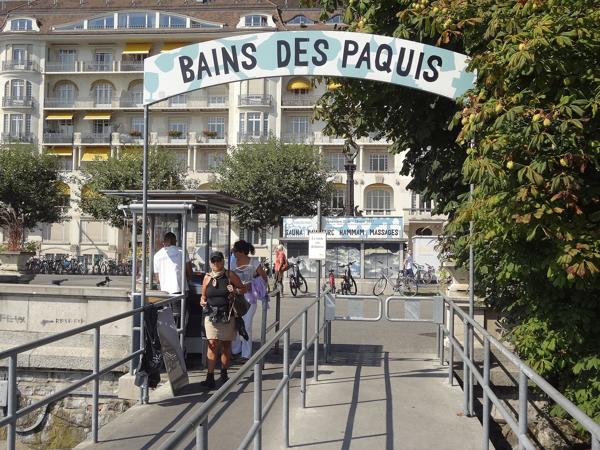 Visitors at the ticket kiosk under the Bains des Pâquis arch on the Quai du Mont-Blanc, with hotels and trees behind
