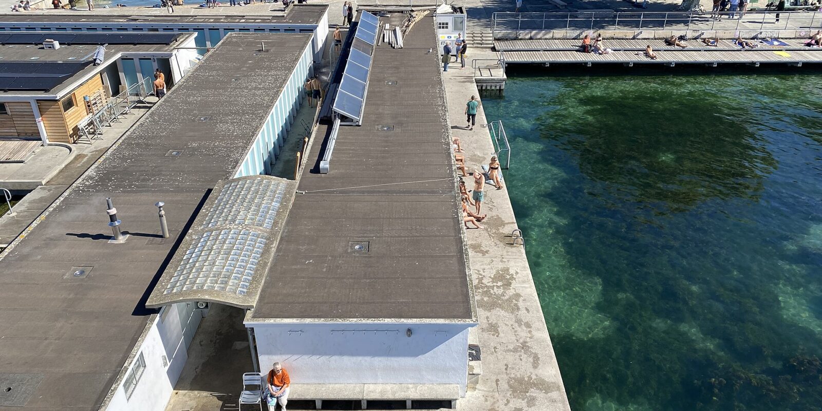 Elevated view over the bath buildings and sun decks of the Bains des Pâquis in Geneva, with bathers beside clear green water