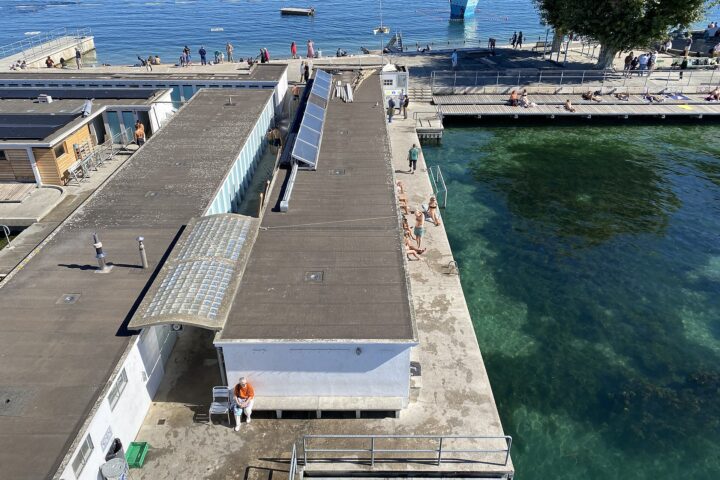 Elevated view over the bath buildings and sun decks of the Bains des Pâquis in Geneva, with bathers beside clear green water