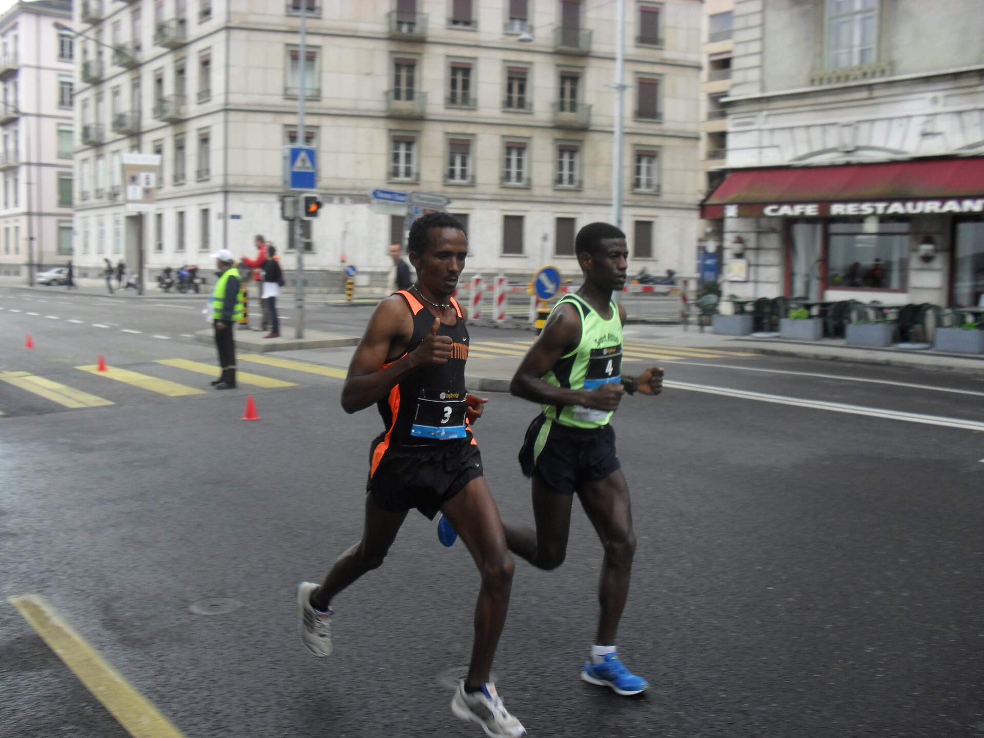 Two leading runners in the Geneva Marathon on a city street, with marshals and a café behind them
