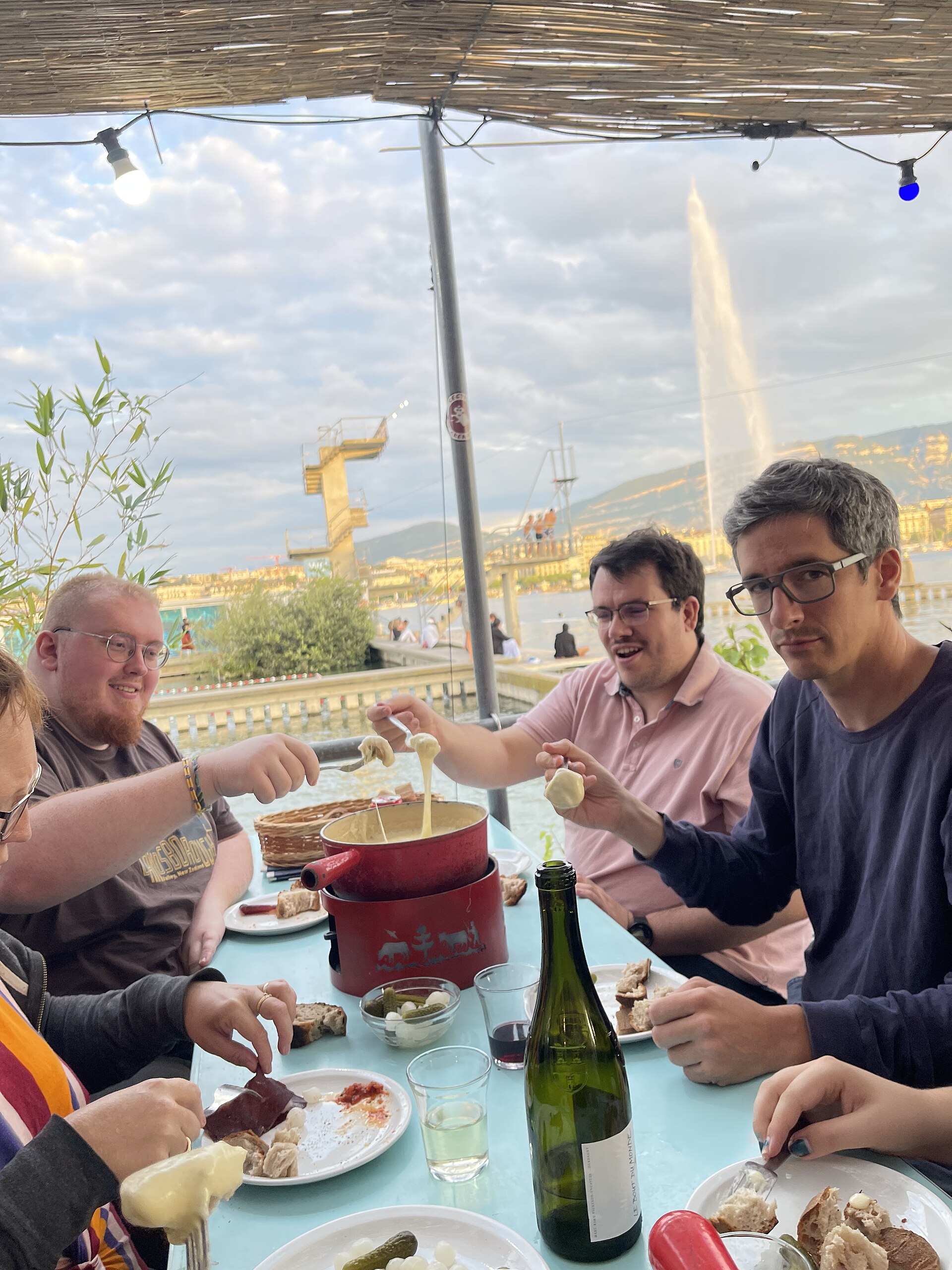 Friends sharing a cheese fondue with white wine at a table at the Bains des Pâquis, with the Jet d'Eau rising across the harbour