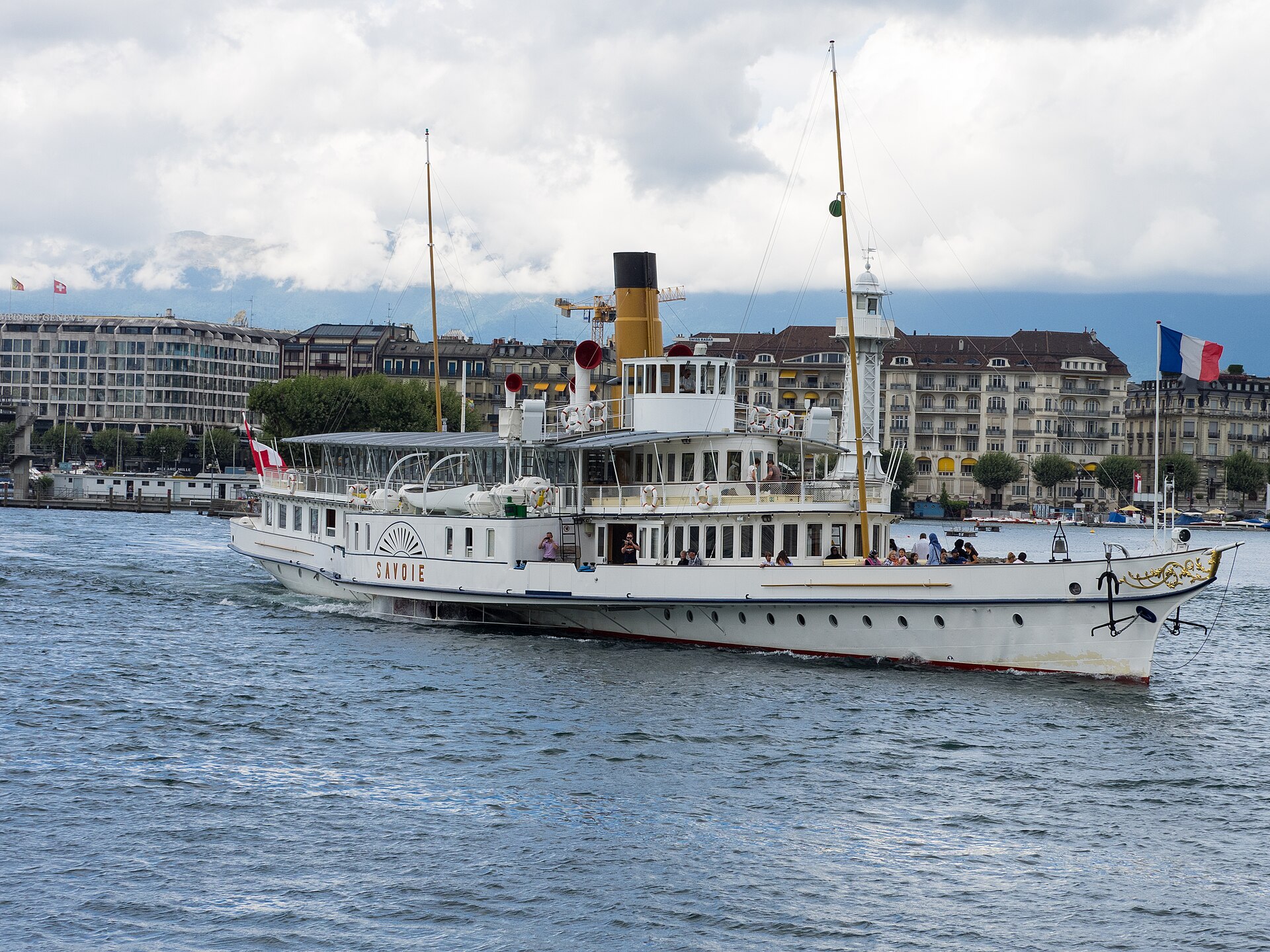 The white paddle steamer Savoie with a yellow funnel sailing in Geneva's harbour, with lakefront buildings behind and a French flag at the bow