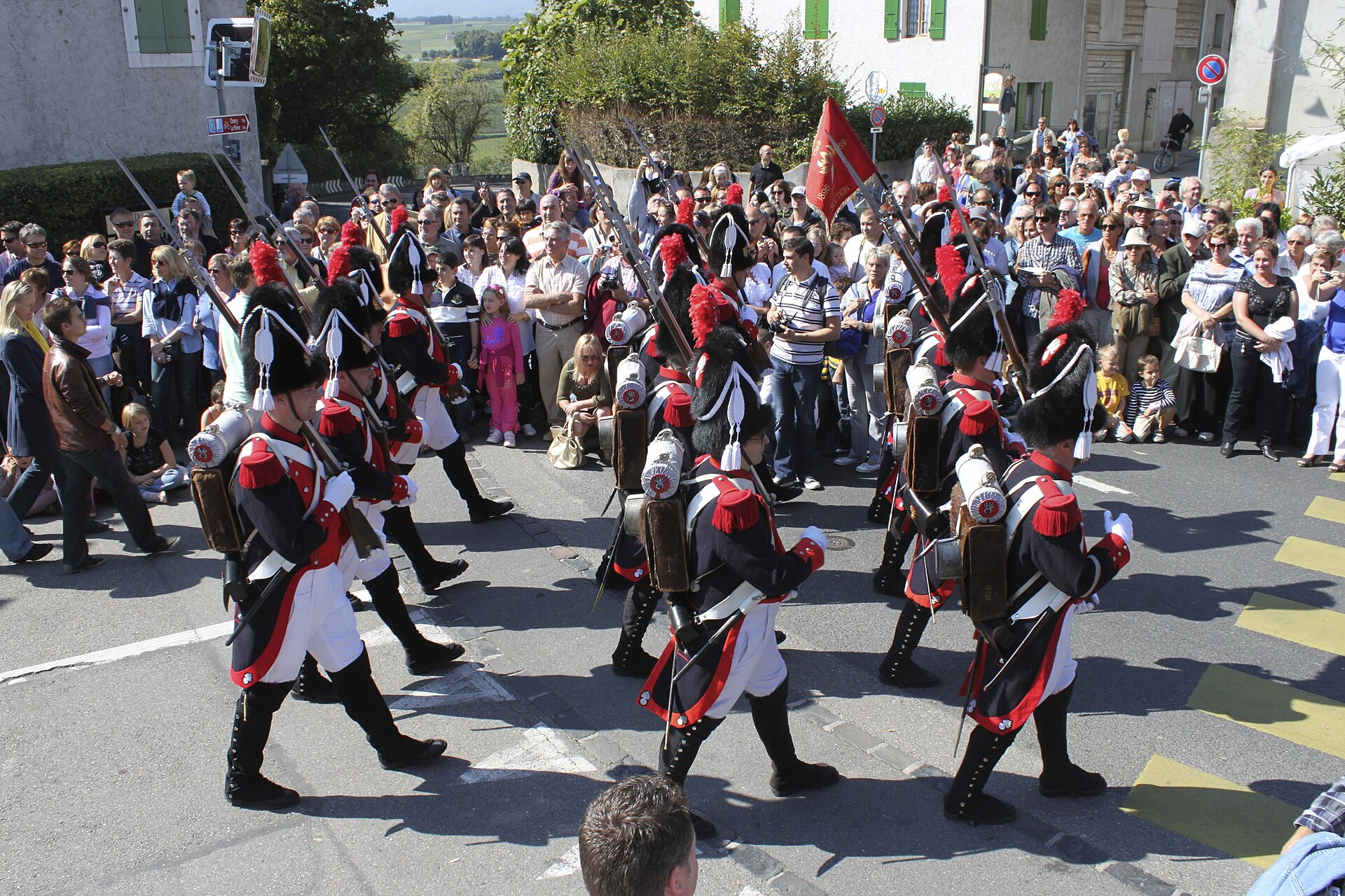 Men in historic red, white and black uniforms with plumed hats marching through a crowd at the Fête des vendanges in Russin