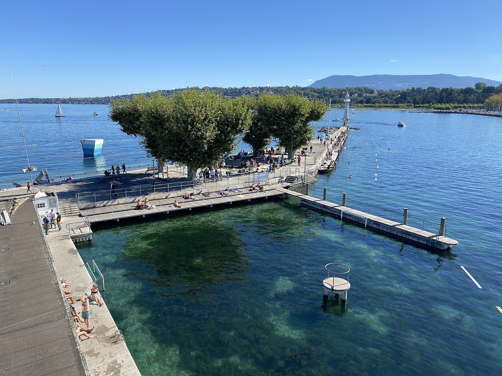 The Bains des Pâquis jetty in Geneva seen from above on a sunny day, with sunbathers on the decks, swimmers in the clear lake and the far shore behind