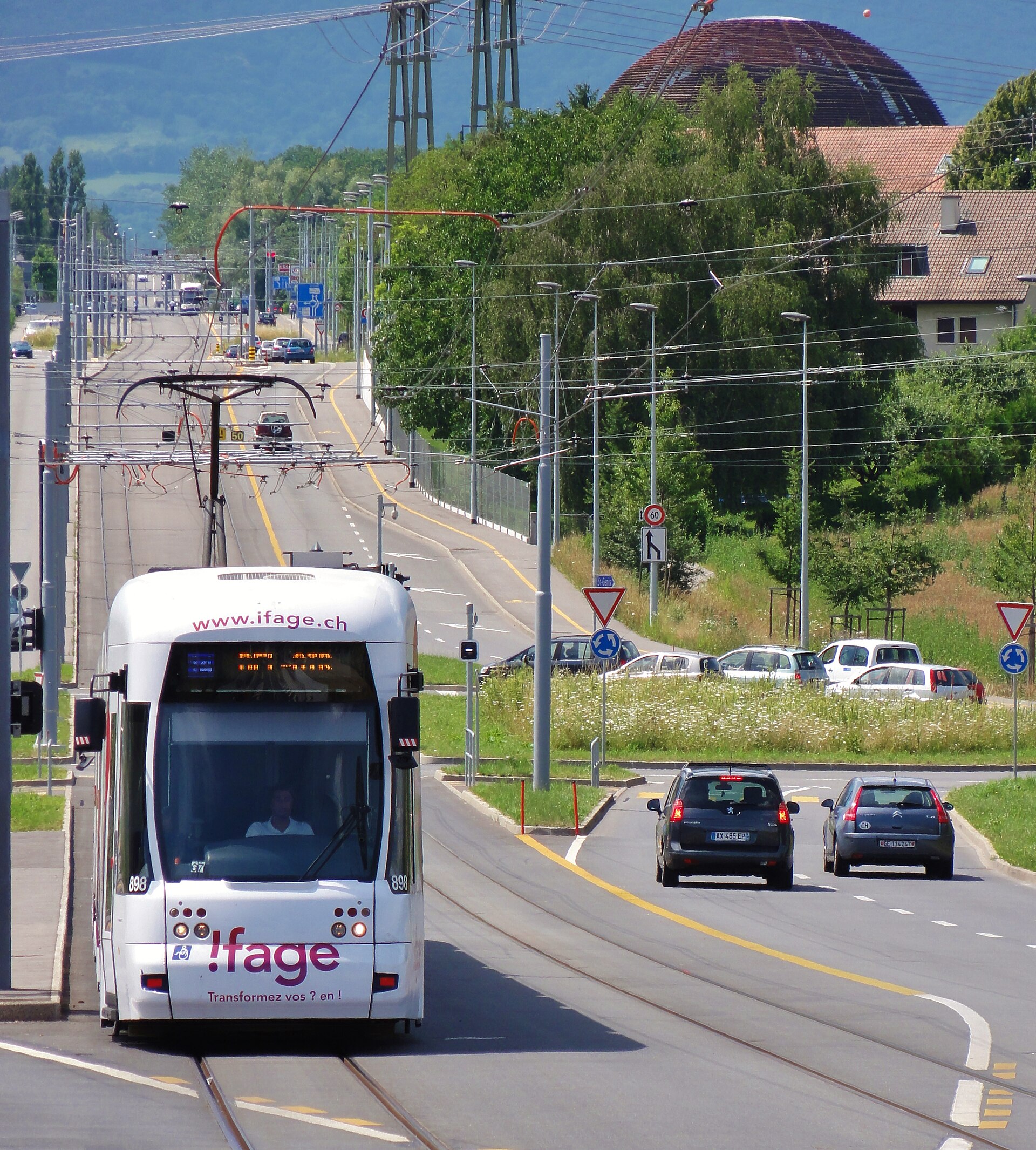 A Geneva line 18 tram approaches along Route de Meyrin, with the CERN Globe visible beyond the overhead wires.