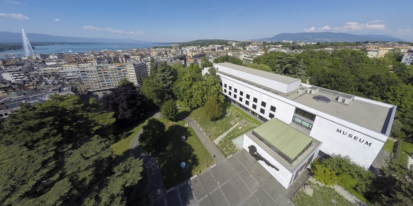 Geneva Natural History Museum seen from above, with parkland, city rooftops, Lake Geneva and the Jet d Eau beyond.