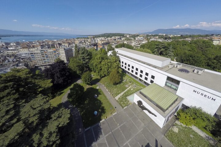 Geneva Natural History Museum seen from above, with parkland, city rooftops, Lake Geneva and the Jet d Eau beyond.