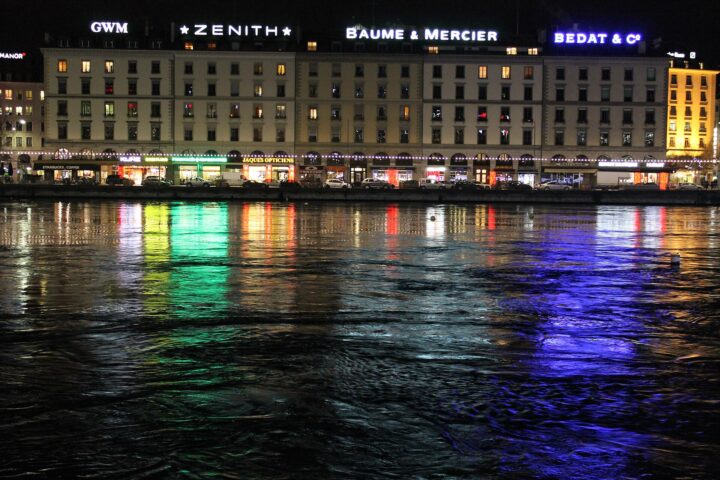 Geneva waterfront at night in December: a row of lit buildings with Zenith, Baume and Mercier and Bedat signs, reflected in green and blue on the water.