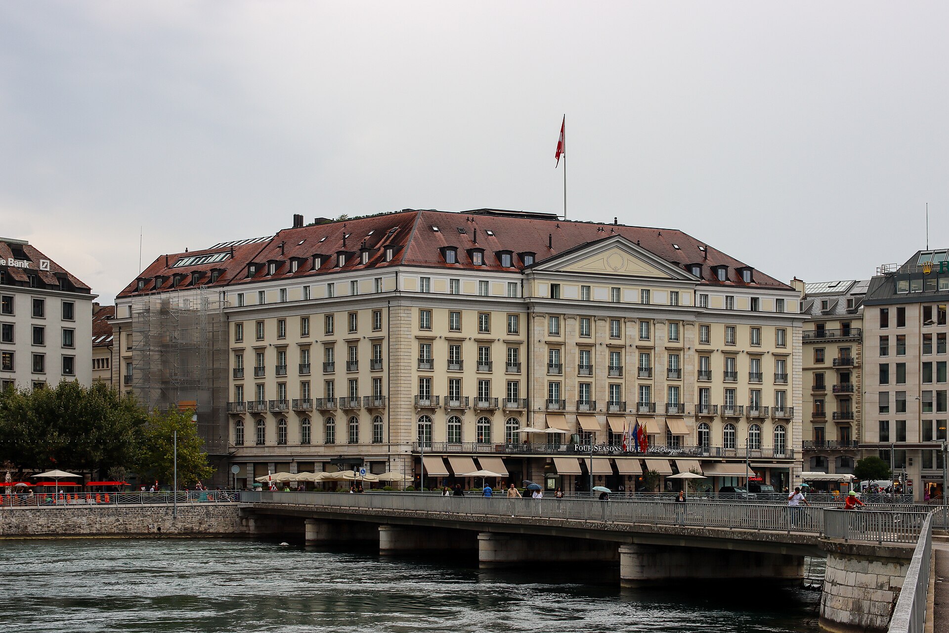 Geneva's Four Seasons Hôtel des Bergues rises behind the pedestrian Pont des Bergues, with river water in the foreground.