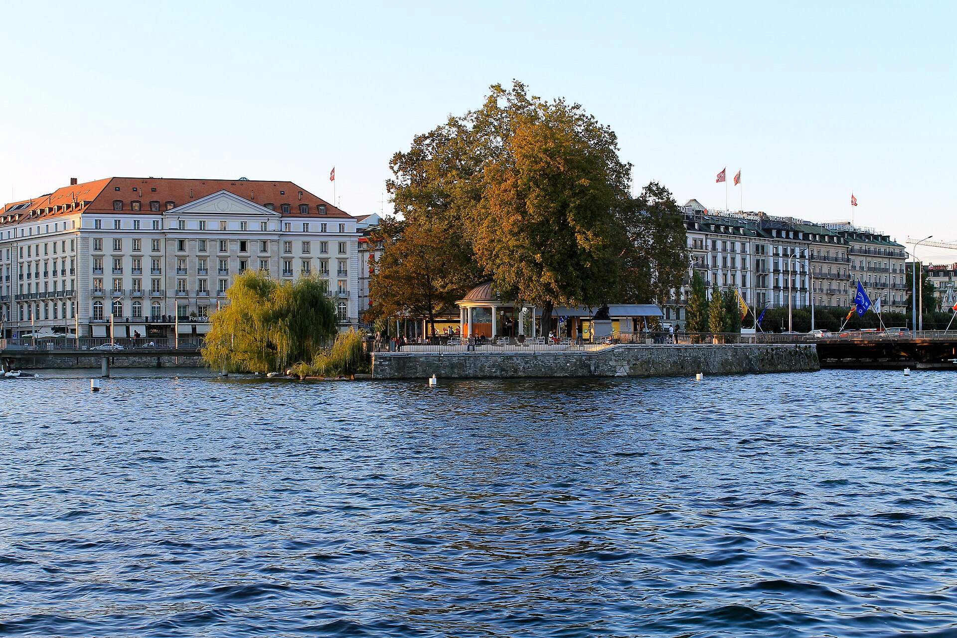 Geneva's Hôtel des Bergues stands beside the waterfront behind Île Rousseau, with trees and a pavilion above blue water.