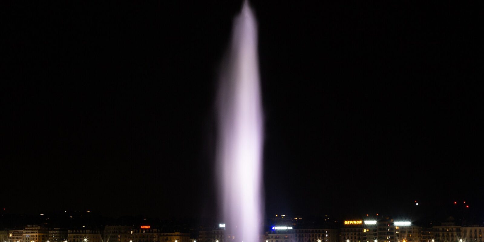 Geneva's illuminated Jet d'Eau rises above dark harbour water, with waterfront lights reflecting beneath the fountain.