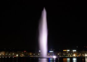 Geneva's illuminated Jet d'Eau rises above dark harbour water, with waterfront lights reflecting beneath the fountain.