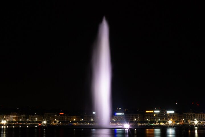 Geneva's illuminated Jet d'Eau rises above dark harbour water, with waterfront lights reflecting beneath the fountain.