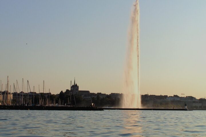 Geneva's Jet d'Eau catches warm evening light beside sailboat masts, with Saint-Pierre Cathedral on the skyline.