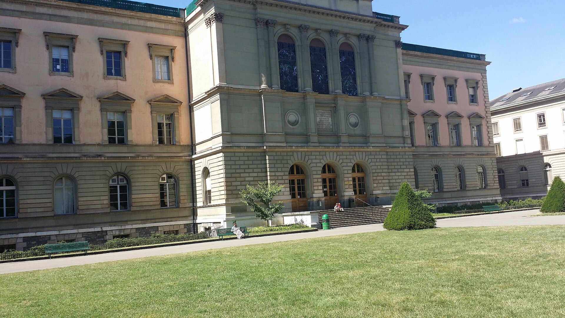 Geneva's Uni Bastions building faces a park lawn and path, with arched doors, tall windows, benches and clipped shrubs.