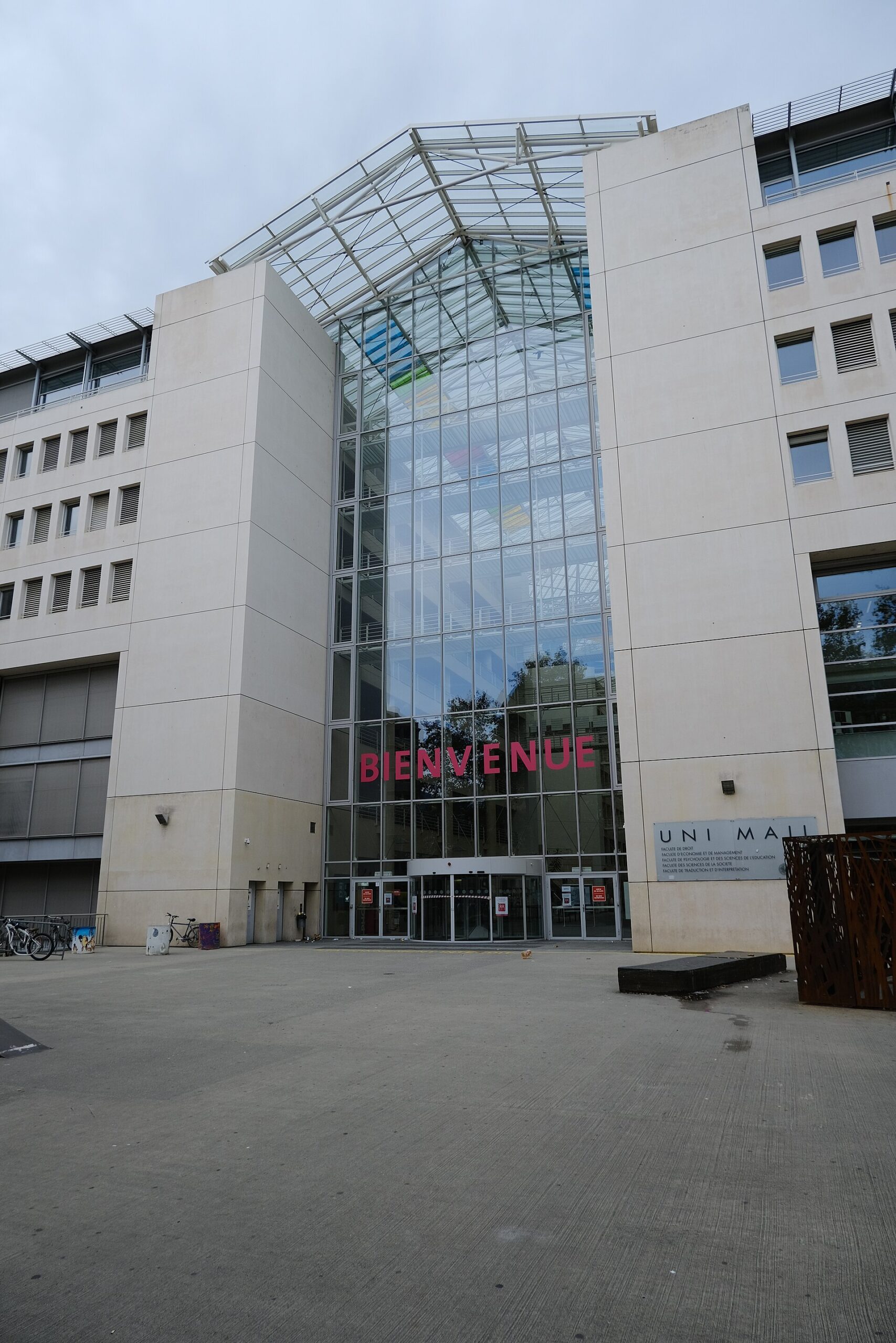 The glass entrance atrium of Uni Mail in Geneva, framed by pale university buildings and an open forecourt.