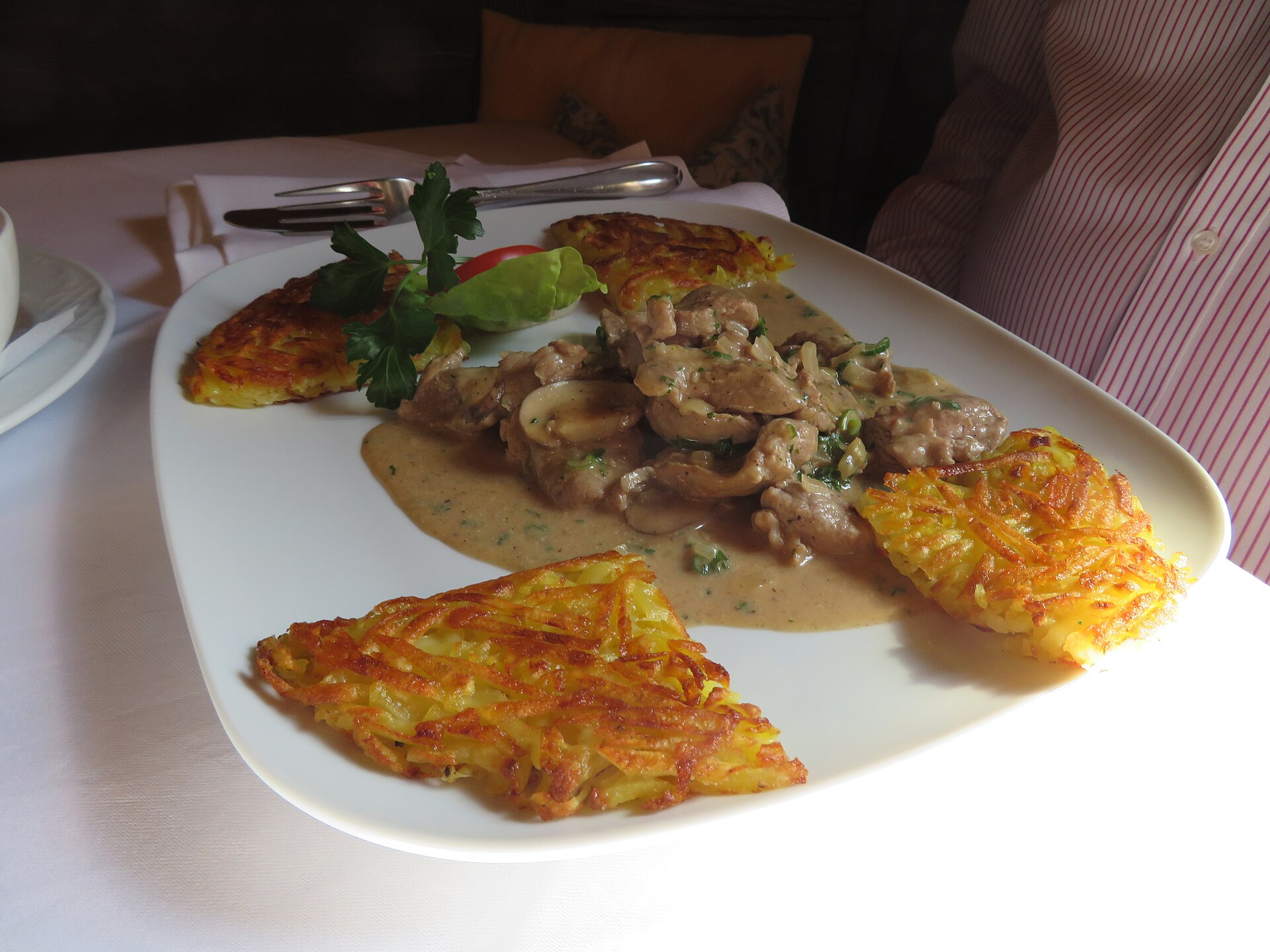 Golden Swiss rösti cakes surround sliced meat and mushrooms in cream sauce on a white restaurant plate.