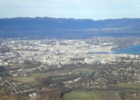 Green fields and the wooded Arve bend lead toward Geneva, Lake Geneva and the Jura in this Salève view.
