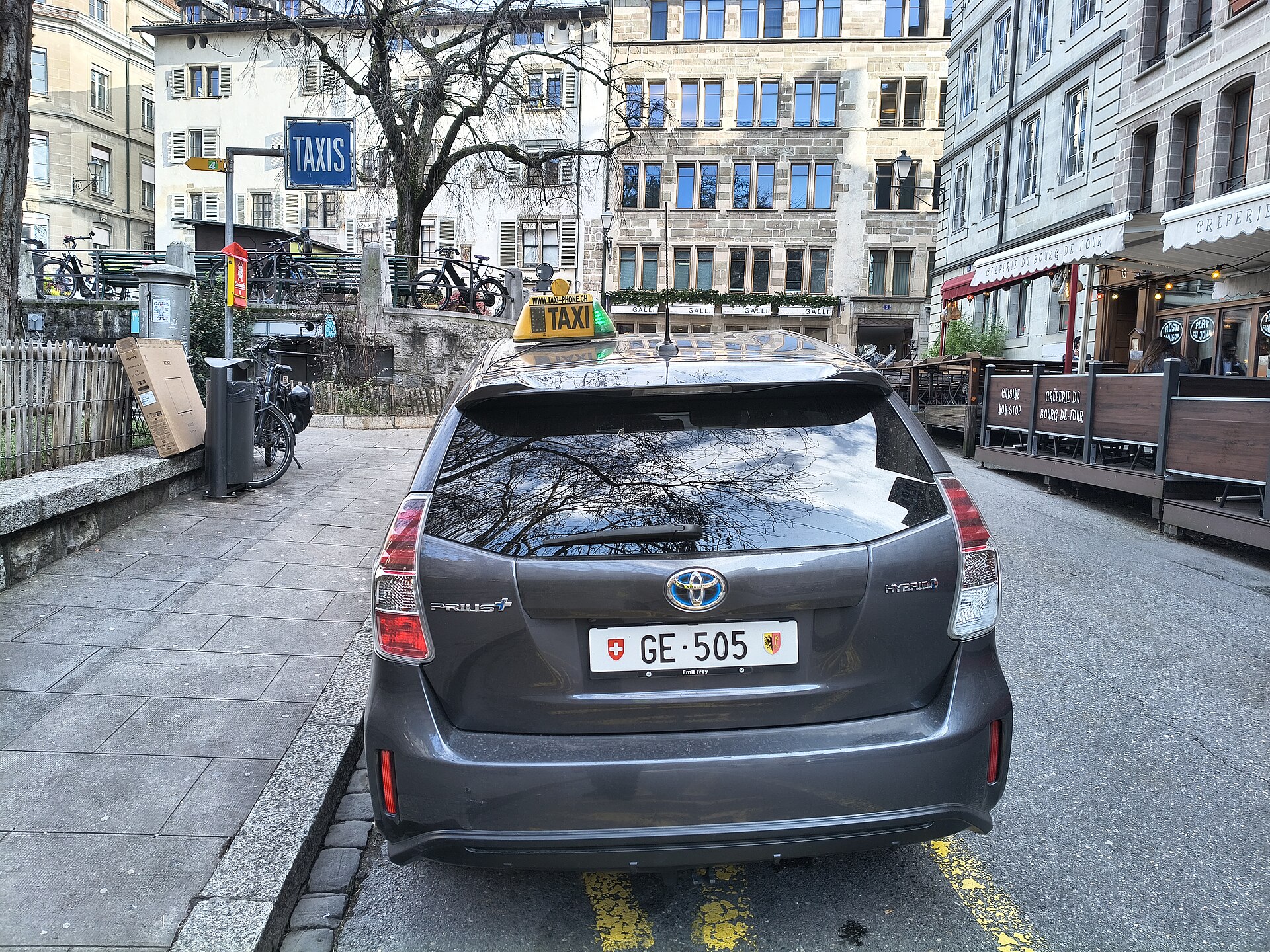 A grey taxi with a yellow roof sign waits in a yellow-marked taxi bay beside a blue TAXIS sign on Place du Bourg-de-Four in Geneva's Old Town.