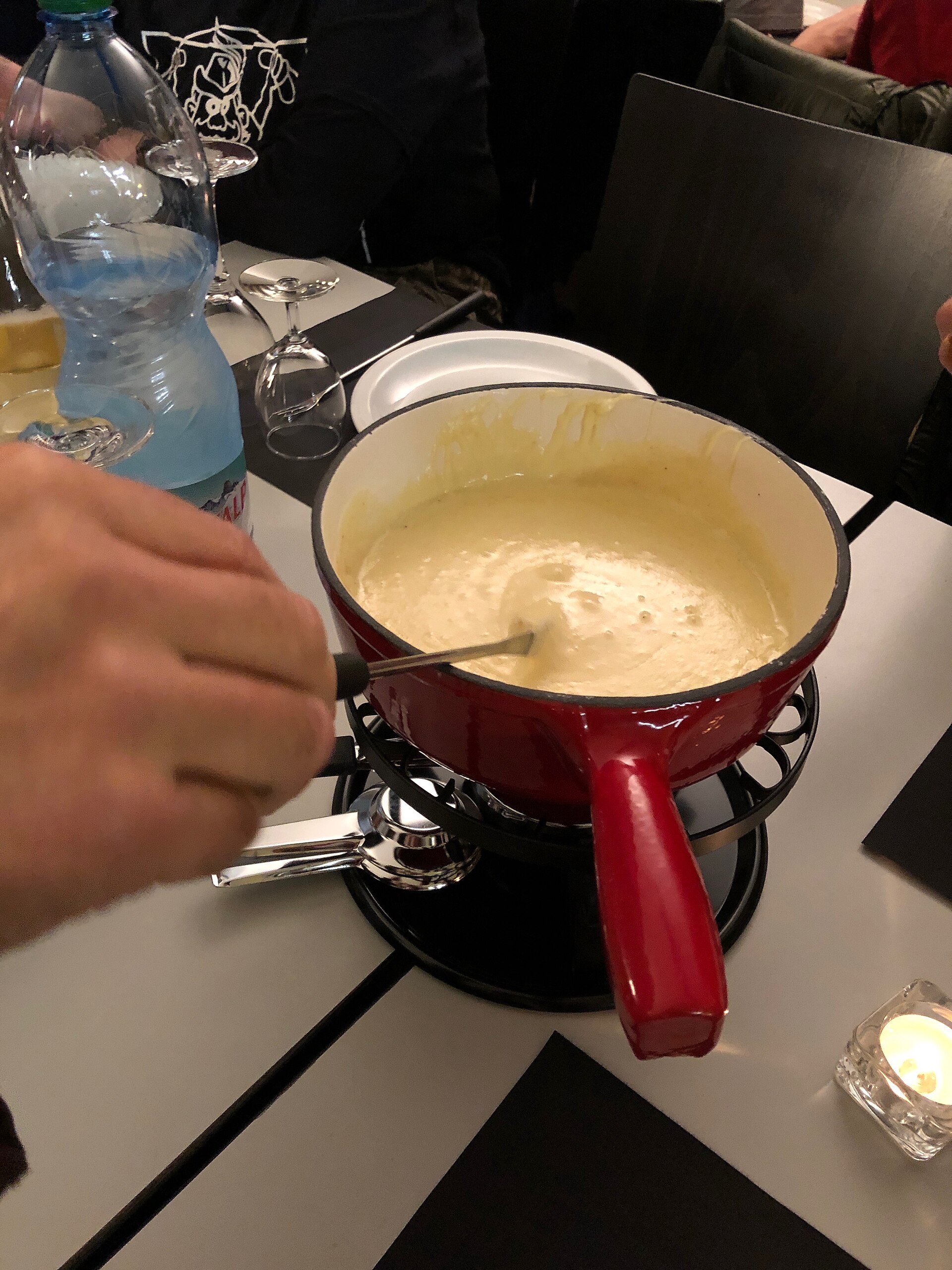 A hand dips a long fork into bubbling Swiss cheese fondue in a red caquelon over a tabletop burner.
