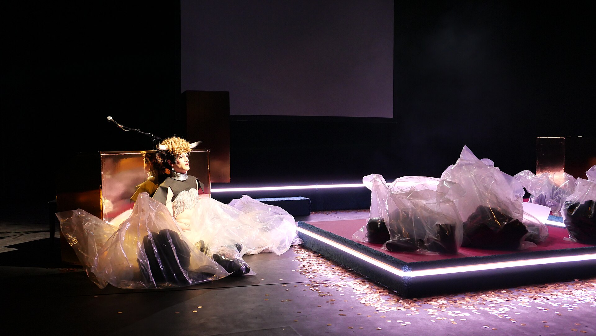 A horned performer sits among translucent materials beside a glowing platform during Dear Human Animals at Geneva’s Antigel festival.