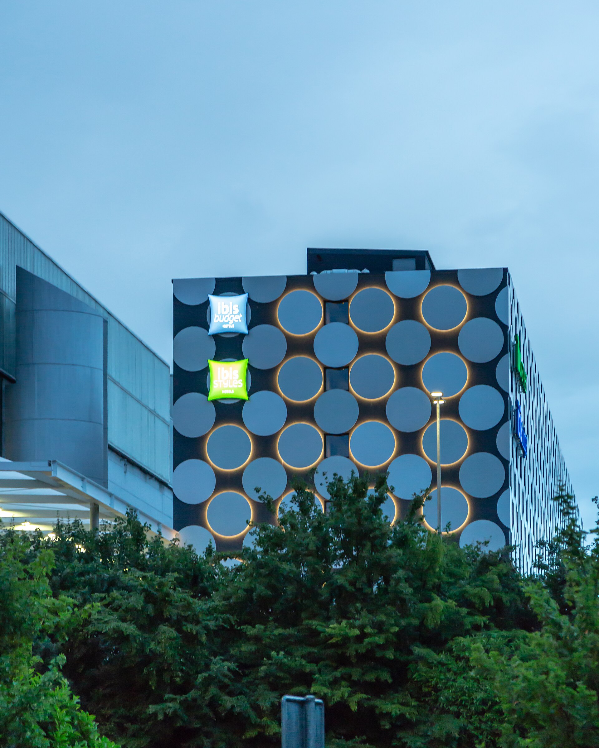 Ibis budget and Ibis Styles signs glow on a circular-patterned hotel facade beside Palexpo, with trees in the foreground.