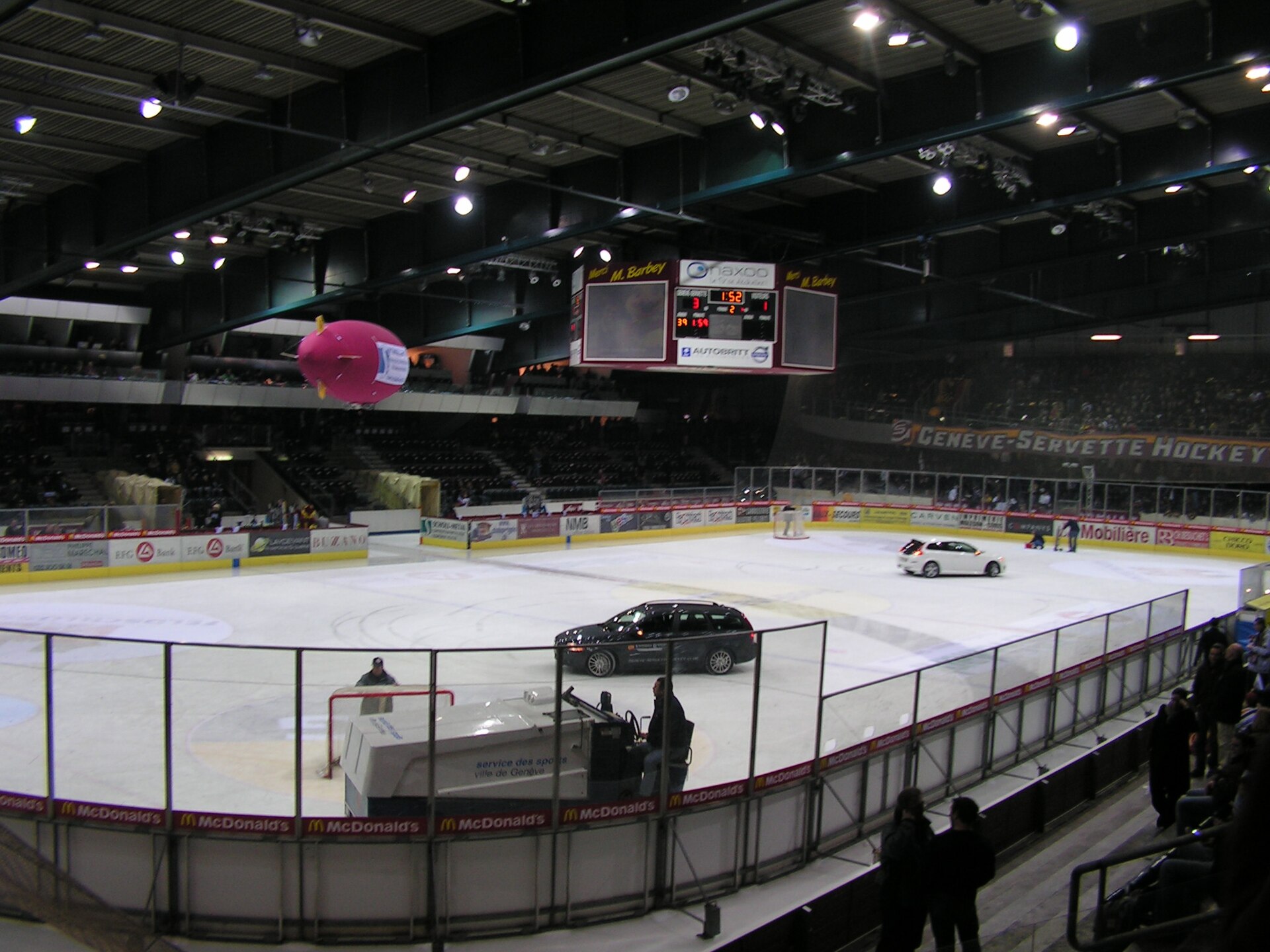 The ice surface of the Patinoire des Vernets in Geneva before a hockey match, with an ice resurfacer, the scoreboard and a Genève-Servette Hockey Club banner above the stands