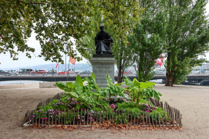 Jean-Jacques Rousseau's seated bronze statue rises above a flower bed on Île Rousseau in Geneva, with a bridge behind.