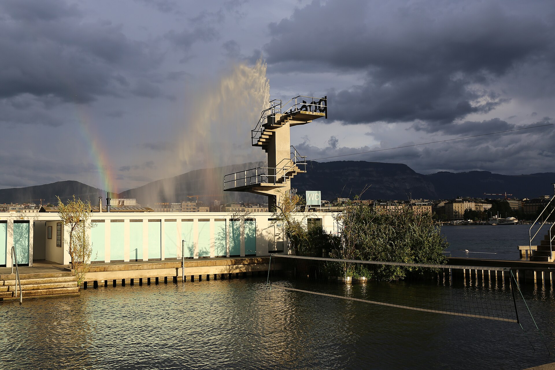 The Jet d'Eau with a rainbow in its spray beyond the diving tower and pavilions of the Bains des Pâquis, under dark clouds