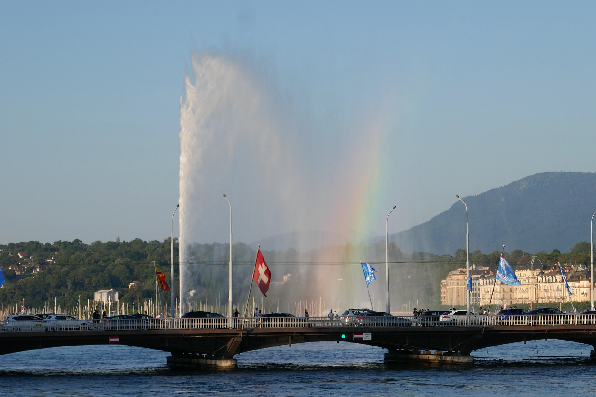 The Jet d'Eau with a rainbow in its spray seen beyond the Pont du Mont-Blanc, with flags along the bridge and a hill behind
