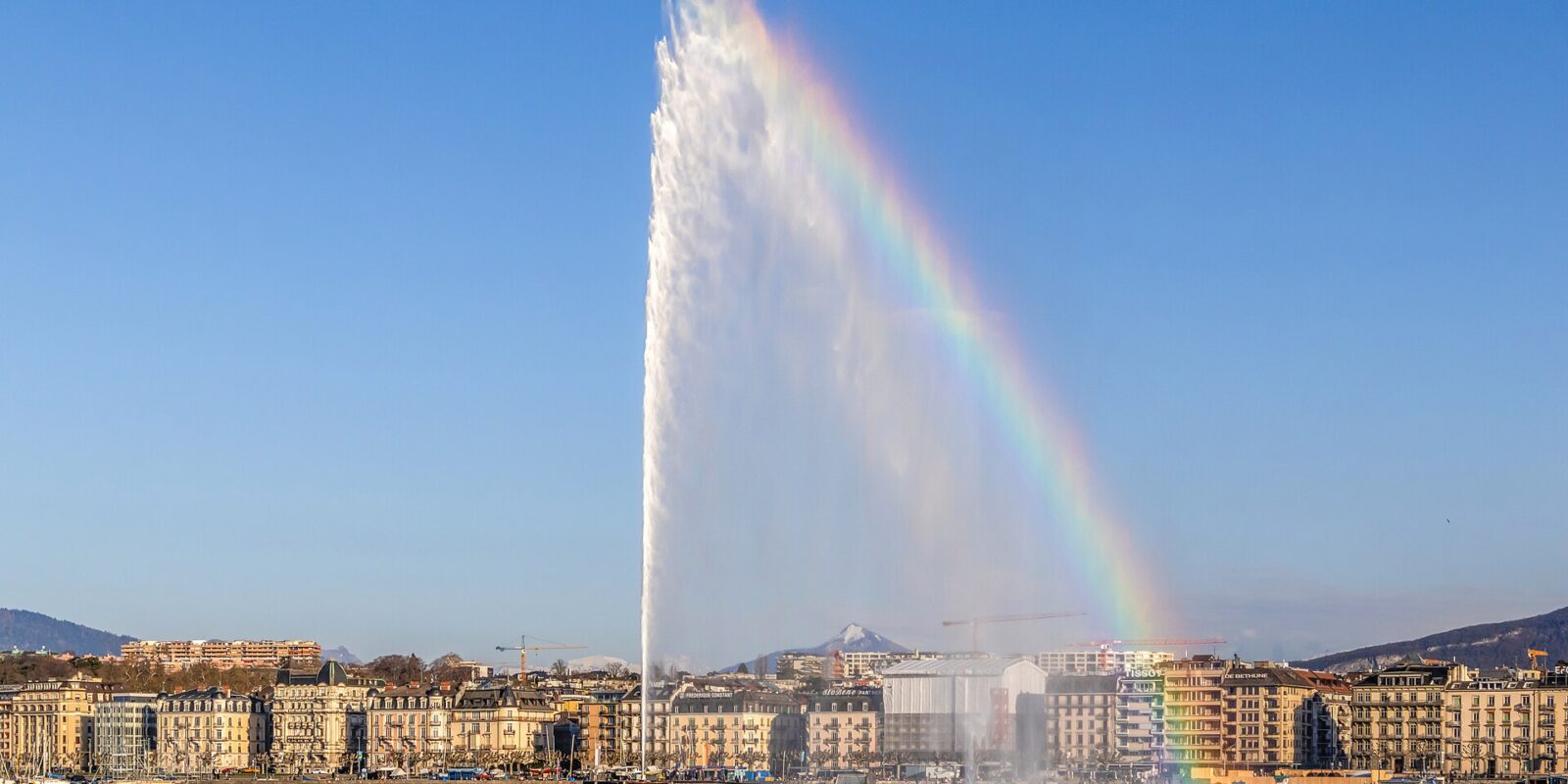 The Jet d'Eau in Geneva shooting high into a blue sky with a rainbow in its spray, above the lake and a row of lakefront buildings