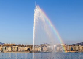 The Jet d'Eau in Geneva shooting high into a blue sky with a rainbow in its spray, above the lake and a row of lakefront buildings