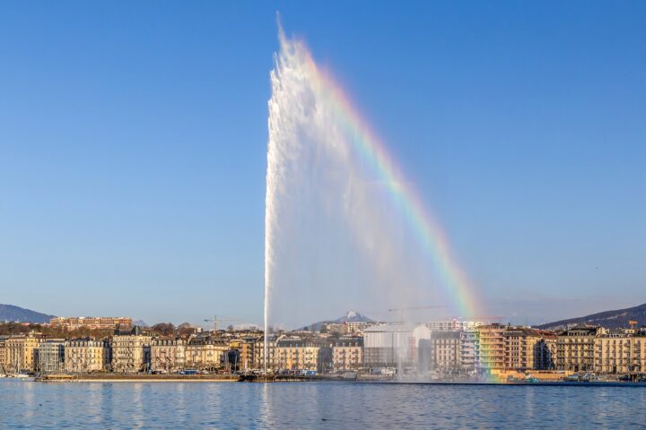 The Jet d'Eau in Geneva shooting high into a blue sky with a rainbow in its spray, above the lake and a row of lakefront buildings