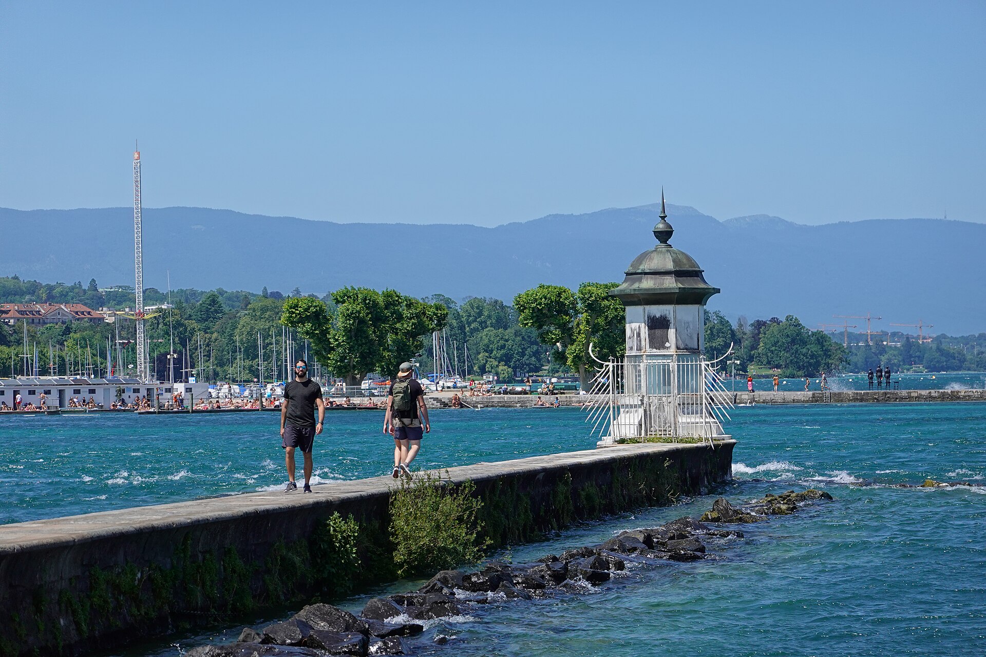 Two people walking along the stone Jetée des Eaux-Vives in Geneva towards its small white lighthouse, with blue lake water and the far shore