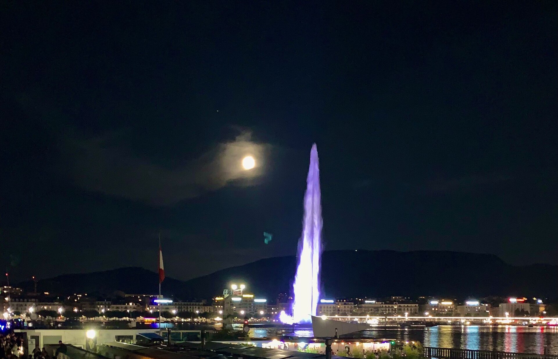 The Jet d'Eau lit in white and violet at night over the dark lake, with city lights along the shore and a full moon above