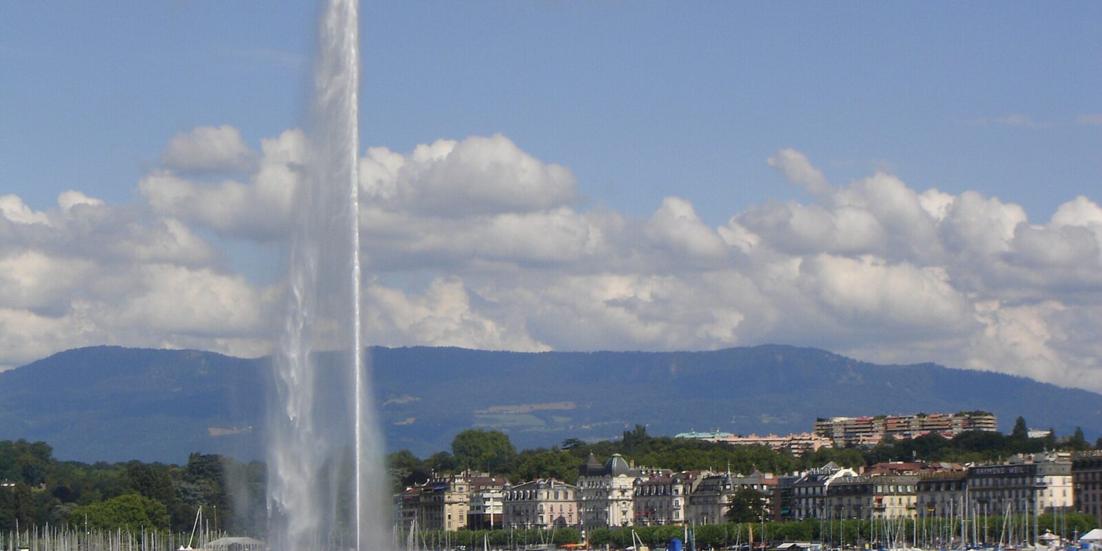 The Jet d'Eau rises beside sailboat masts and waterfront buildings across Geneva harbour under scattered clouds.