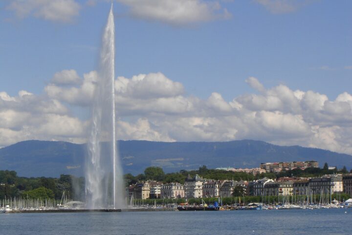 The Jet d'Eau rises beside sailboat masts and waterfront buildings across Geneva harbour under scattered clouds.
