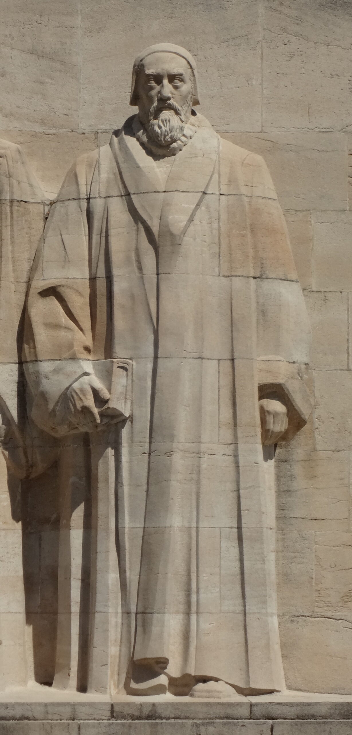 John Knox's full-length stone statue holds a book against the pale stone blocks of Geneva's Reformation Wall.