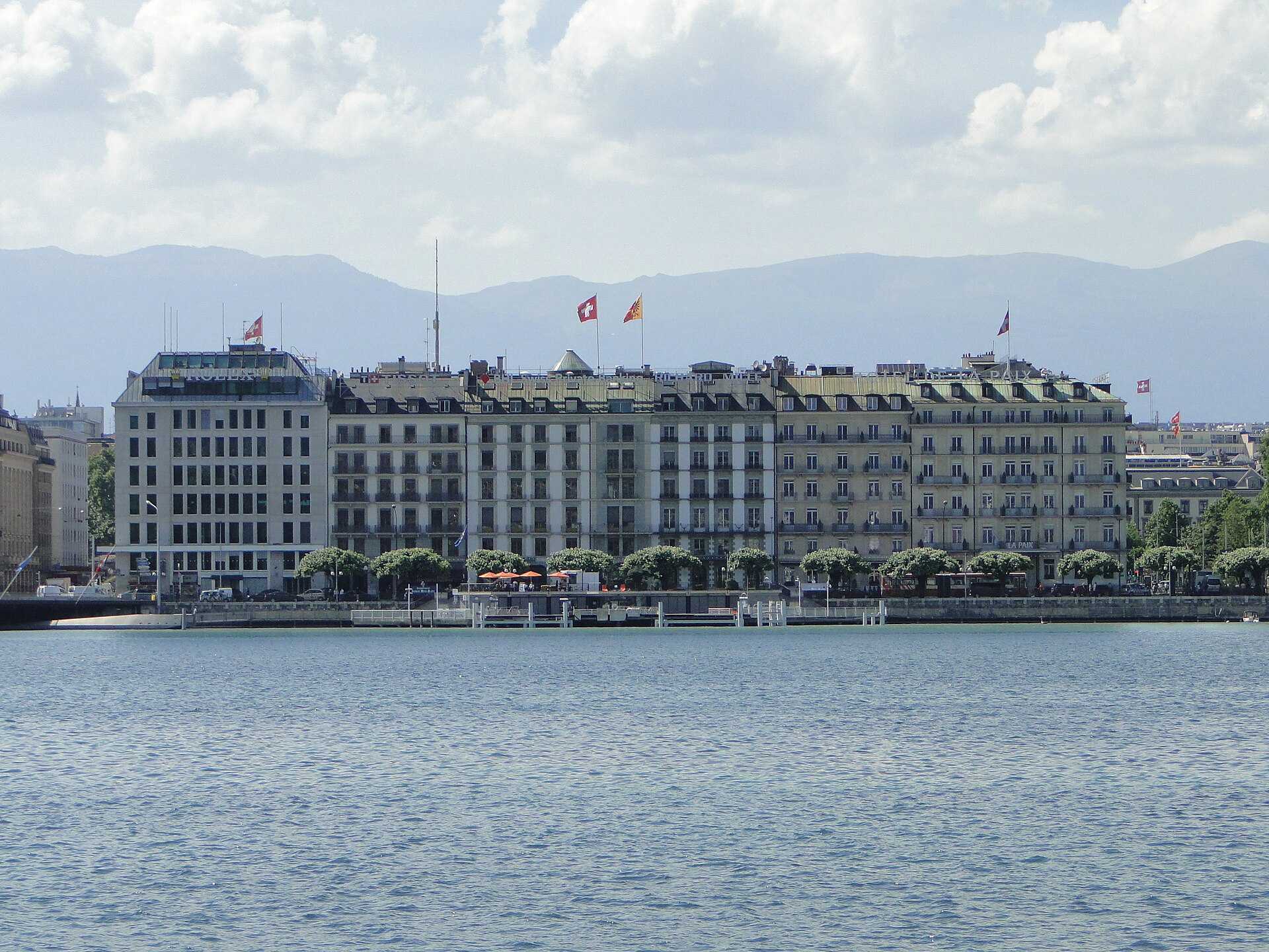 Lake Geneva faces the grand hotel facades of Quai du Mont-Blanc, with rooftop signs and flags above the waterfront trees.