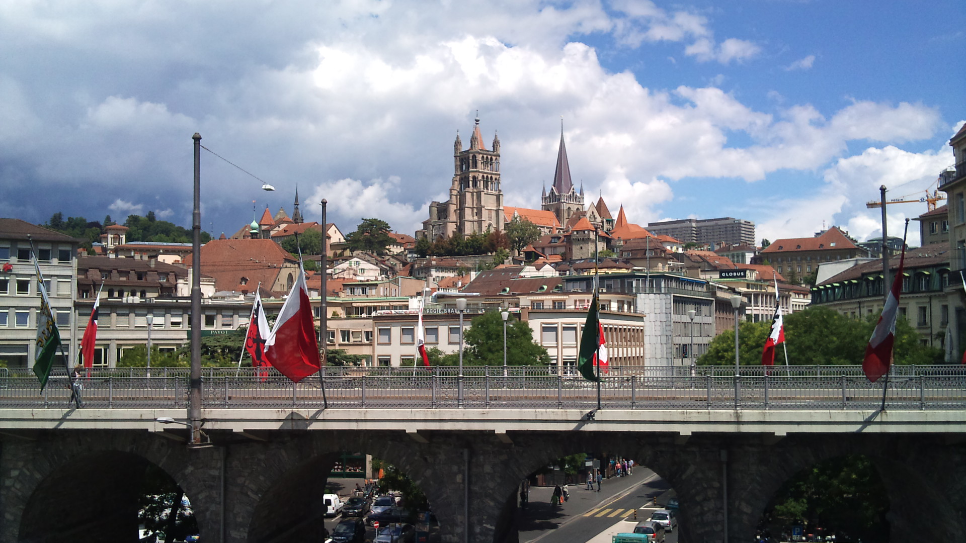 Lausanne Cathedral rises above red-roofed buildings beyond the flag-lined Grand Pont under a sky of large clouds.
