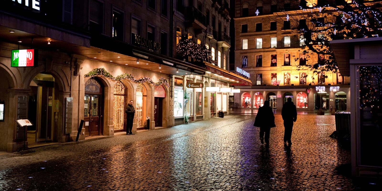 Lit restaurant and pub fronts on a wet cobbled square in Geneva at night, with festive lights in a tree and two people walking across