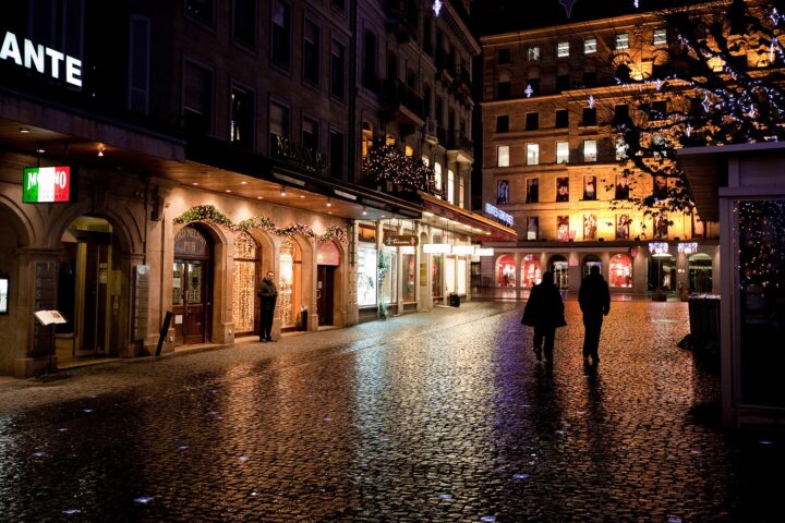 Lit restaurant and pub fronts on a wet cobbled square in Geneva at night, with festive lights in a tree and two people walking across