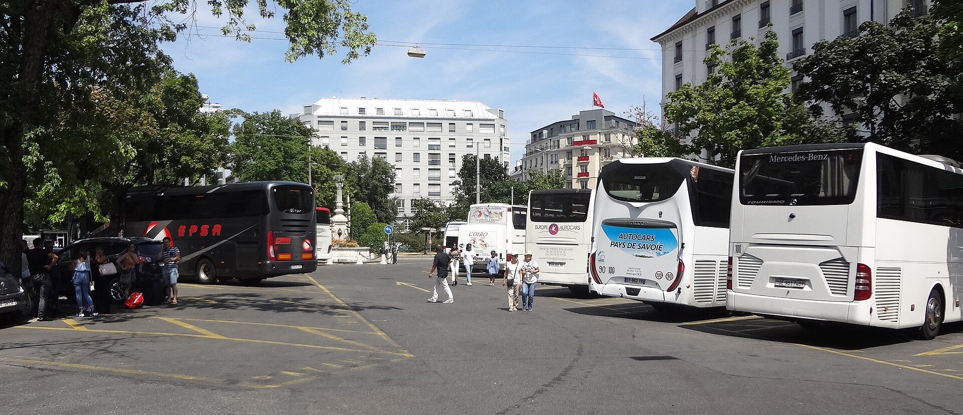 Long-distance coaches stand in marked bays at Geneva's Place Dorcière bus station, with passengers and luggage beside the vehicles.