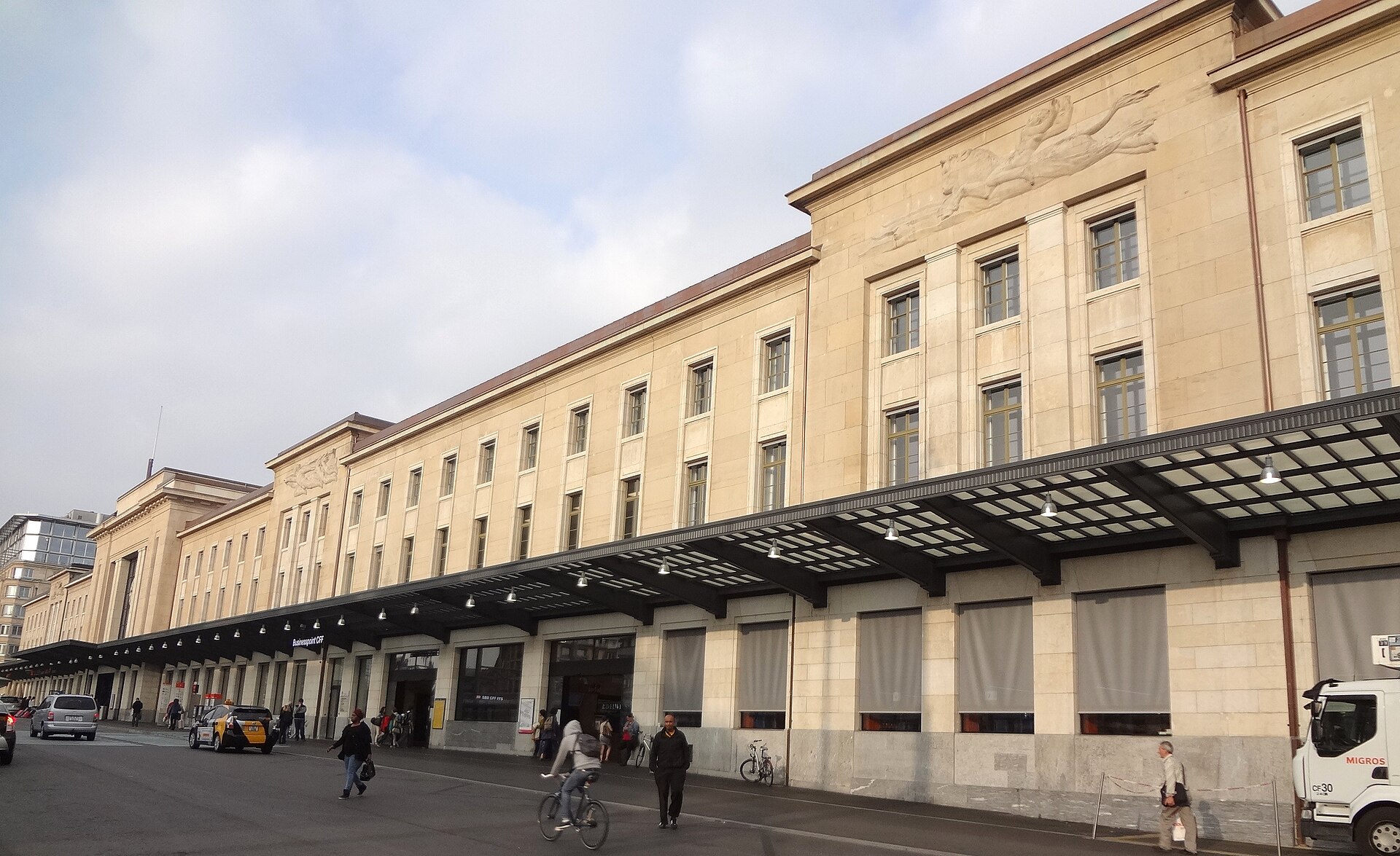 The long stone facade of Geneva Cornavin station stretches beside a covered pavement with pedestrians and a passing cyclist.