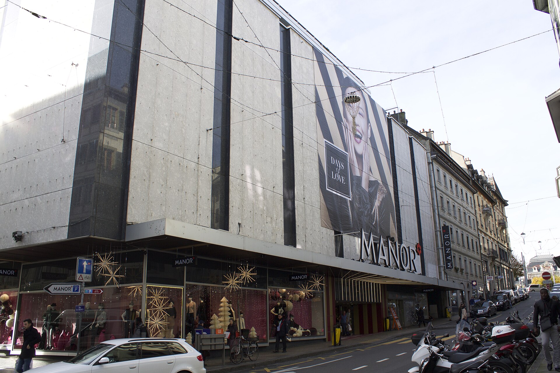 Manor's department store facade and display windows rise above pedestrians and parked motorcycles in central Geneva.
