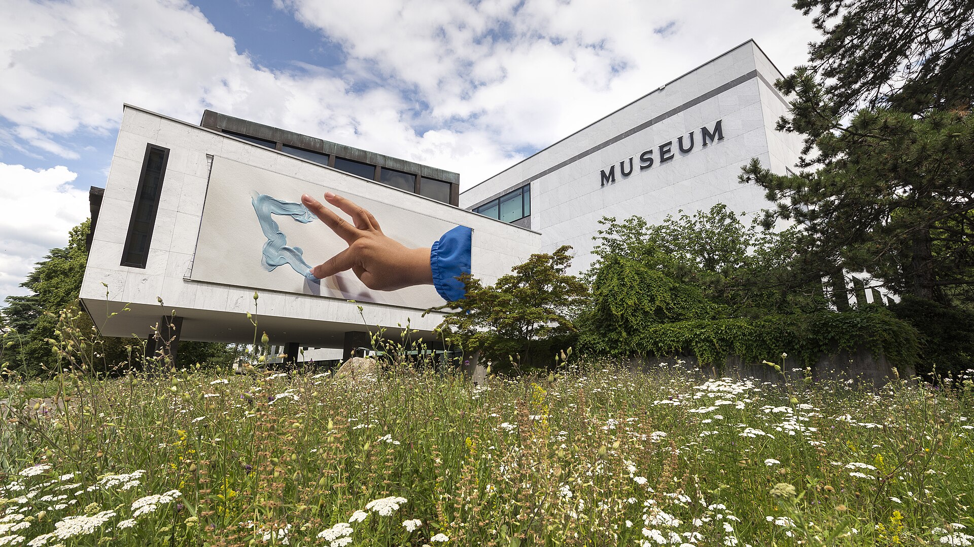 Meadow flowers grow before the white Geneva Natural History Museum, whose projecting entrance carries a large illustrated banner.